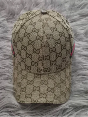 Beige keps från Gucci med GG-mönster - Snygg beige keps från Gucci med klassiskt GG-monogram över hela kepsen. Kepsen har böjd skärm och en grön-röd rand på sidan som ger en extra touch. Tillverkad i canvasmaterial som känns både lyxigt och hållbart. Perfekt för dig som vill sticka ut med en ikonisk accessoar.