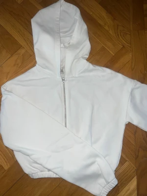 Vit zip hoodie - Snygg vit hoodie med dragkedja framtill och huva. Modellen har långa ärmar och resår i midjan samt vid ärmsluten. Perfekt för en clean och enkel stil. Materialet känns mjukt och bekvämt mot huden.