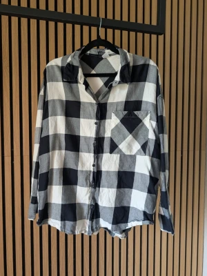Svartvit rutig skjorta i bomull - Säljer en oversized svartvit rutig skjorta med klassisk krage och knappstängning framtill. Skjortan har en bröstficka med diagonalt mönster och långa ärmar. Perfekt för en avslappnad och trendig look.