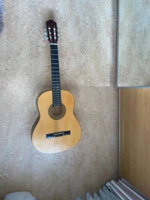 Klassisk gitarr från Cataluna - Säljer en klassisk akustisk gitarr från Cataluna med naturfärgad kropp i trä och dekorativ rosett runt ljudhålet. Gitarren har sex nylonsträngar och mörkbrunt huvud. Perfekt för dig som vill börja spela eller bara jamma hemma.