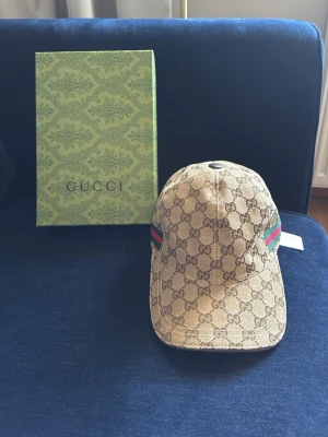Gucci keps med monogram och ränder - Snygg beige keps från Gucci med klassiskt GG-monogram över hela kepsen och gröna/röda ränder på sidorna. Kepsen har böjd skärm och justerbar passform. Materialet är canvas med detaljer i läder. Perfekt för dig som gillar lyxiga accessoarer.