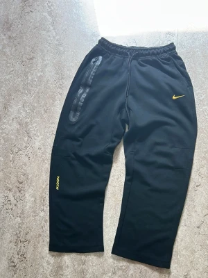 Svart Nike tech NOCTA  byxor - Svart loungewear set från Nike NOCTA med matchande hoodie och byxor. Setet har gula detaljer, inklusive NOCTA- och Nike-loggor. Hoodien har dragkedja och fickor med svarta dragkedjor. Byxorna har resår i midjan och en liten gul Nike-logga. Byxorna står det st M fast passar som S och samma sak med Nike tech koftan så står det st S men passar Xs. Pris går diskutera 