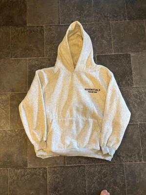 Grå Essentials Fear of God hoodie - Snygg grå hoodie från Fear of God Essentials med svart logga på bröstet. Klassisk passform med huva och känguruficka framtill. Mjuk bomullsblandning och ribbade muddar vid ärmslut och nederkant. Perfekt för en avslappnad streetwear-look.