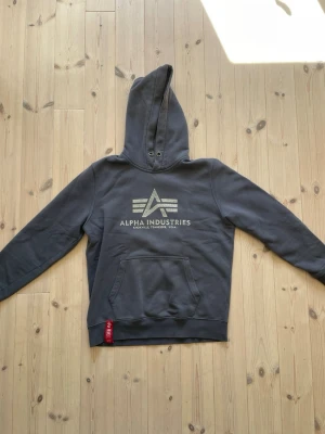 Mörkgrå hoodie Alpha Industries - Mörkgrå hoodie från Alpha Industries med stor logga och text på bröstet. Klassisk känguruficka framtill och en röd tagg med logga vid nederkanten. Hoodien har huva med metallöljetter och är gjord i mjukt bomullsmaterial. I hyfsat skick. 