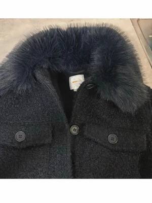 Gina  - Stilren svart kappa från Ganni med fluffig mörkblå fuskpäls på kragen. Kappan har två bröstfickor med knappar och en grov, texturerad yta. Perfekt för dig som vill ha en trendig och varm look i vinter.