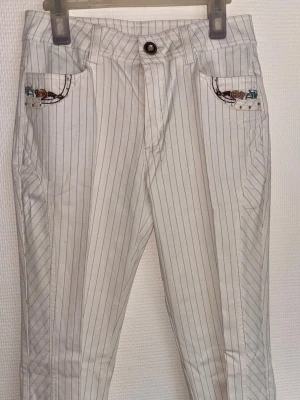Vita randiga jeans med broderi - Snygga vita jeans med tunna vertikala ränder och unika broderade detaljer på fickorna. Byxorna har guldfärgade nitar och klassisk femficksmodell. Perfekta för dig som vill sticka ut med en cool och personlig stil. Made in korea. Alla knappar finns kvar, finns nån fläck som går bort i tvätten. Använd fåtal gånger. 