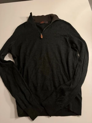 Mörkgrå stickad tröja med halvzip från Morris - Snygg mörkgrå stickad tröja från Morris med halvzip och hög krage. Tröjan har långa ärmar, ribbade muddar och en diskret logga på bröstet. Perfekt för lager-på-lager och passar till det mesta.