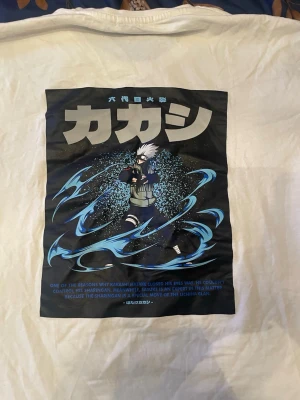 Vit anime t-shirt med Kakashi-tryck - Vit t-shirt med stort färgglatt tryck av Kakashi från Naruto på framsidan. Trycket har japansk text och coola blå effekter runt karaktären. T-shirten är i bomull och har klassisk passform med korta ärmar. Perfekt för animefans!