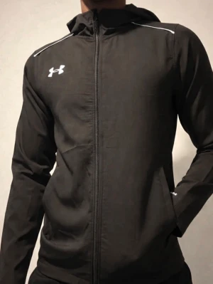Svart träningsjacka Under Armour - Svart träningsjacka från Under Armour med huva och hel dragkedja. Jackan har vita detaljer på axlarna, logga på bröstet och stort Under Armour-tryck på ryggen. Tillverkad i lätt och funktionellt material, perfekt för träning eller uppvärmning.
