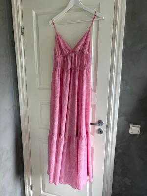 Rosa långklänning med zebramönster - Luftig rosa långklänning med tunna axelband och v-ringning. Klänningen har ett subtilt zebramönster i ljusrosa toner och är tillverkad i ett lätt, somrigt tyg som faller snyggt. Perfekt för varma dagar och ger en avslappnad men trendig look.