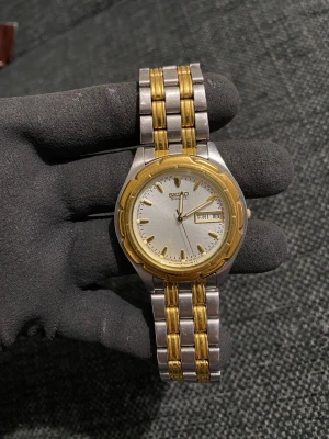 Seiko Quartz two-tone watch (7N43-7071) - Säljer en klassisk Seiko Quartz-herrklocka (modell 7N43-7071) i tvåfärgat utförande. Klockan har dag- och datumvisning.  Klockan är ej funktionstestad och säljes i befintligt skick. Batteriet har dött. Det finns normalt bruksslitage pả boett och länk, samt lite synliga repor på glaset (se bilder för egen bedömning).  Länken passar just nu en handled pả ca 16,5 cm. Det går att förlänga länken något (några millimeter) genom att flytta sprintarna/pinnarna i spännets mikrojustering. 
