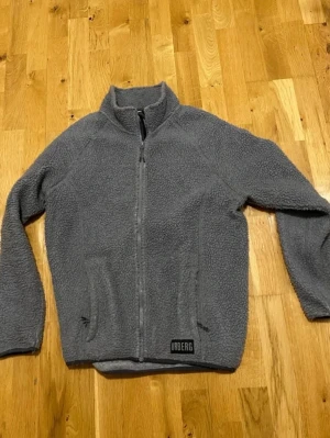 Grå teddyjacka från Urberg stl M - Mysig grå fleece från Urberg med hel dragkedja framtill och hög krage. Jackan har två fickor med dragkedja och en klassisk, rak passform. Perfekt för kyliga dagar när du vill vara både varm och stilren.