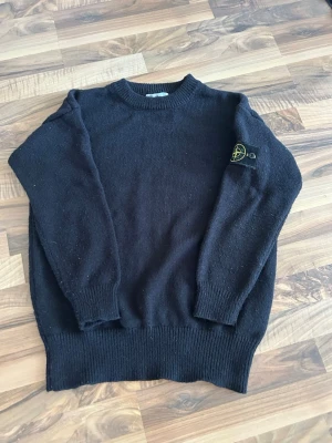 Svart stickad tröja från Stone Island - Svart stickad tröja från Stone Island med klassisk rund halsringning och ribbade muddar. Tröjan har den ikoniska Stone Island-loggan på vänster ärm. Perfekt för dig som gillar streetwear och stilren design.