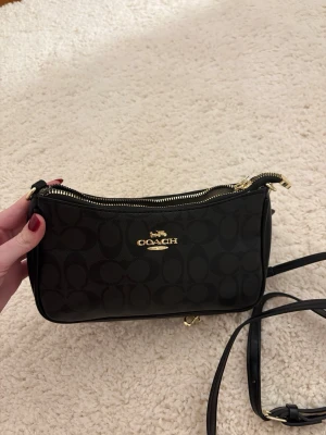 Svart axelremsväska från Coach - Snygg svart handväska från Coach med klassiskt monogrammönster och guldfärgade detaljer. Väskan har en smidig axelrem och dragkedja upptill. Perfekt storlek för det viktigaste och en stilren look med lyxig känsla.