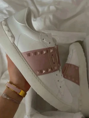 Valentino Opens - Snygga vita sneakers från Valentino med bred rosa rem över mitten, dekorerad med ikoniska pyramidformade nitar. Skorna är i läder och har en chunky vit sula samt nitar även baktill. Perfekta för dig som gillar en stilren men edgy look.Og box och dustbag följer med 😊