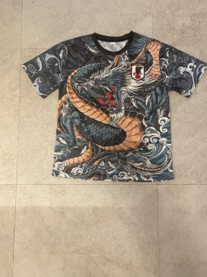 Japan fotbollströja med drakmönster och badge - Cool t-shirt med stort drakmotiv i blått, beige och svart. Badge med japansk stil på bröstet. Kortärmad modell med rund hals. Materialet känns som polyester, perfekt för en unik streetstyle.