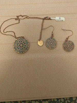 Set med halsband och örhängen Guld Sverige - Snyggt smyckeset från Guld Sverige med halsband och matchande örhängen. Båda har runda hängen i guldfärgad metall med turkosblått, detaljerat mönster i orientalisk stil. Perfekt för dig som gillar unika accessoarer med boho-vibe.