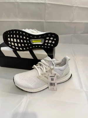 Vita Adidas Ultraboost sneakers - Fräscha vita Adidas Ultraboost sneakers med stickad ovandel i mesh, klassiska tre ränder på sidan och svart sula med Boost-dämpning. Skorna har vita snören och detaljer i ljusgrått. Perfekta för dig som gillar stilrena och bekväma sneakers.