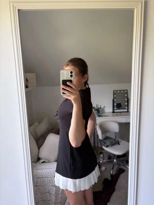 Svart klänning med vit plisserad kant - Snygg svart kortklänning med vit plisserad volangkant nertill. Klänningen har en figurnära passform och korta ärmar. Perfekt för dig som gillar en stilren look med en twist. Ingen synlig logga eller märke.