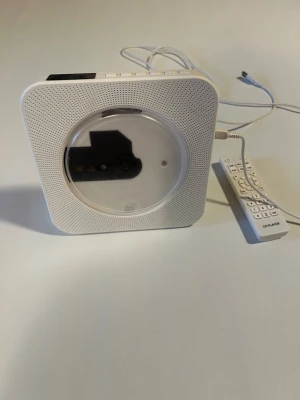 Vit CD-spelare med fjärrkontroll - Säljer en stilren, fyrkantig CD-spelare i vit plast med rund öppning i mitten och tillhörande fjärrkontroll. Spelaren har inbyggda högtalare och USB-kabel för ström. Perfekt för att spela CD-skivor hemma eller på rummet. 