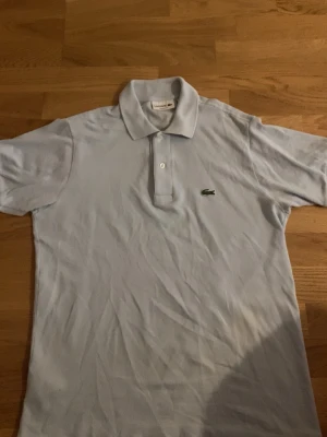  pikétröja från Lacoste - Säljer en klassisk ljusblå pikétröja från Lacoste med krage och två knappar framtill. Tröjan har korta ärmar och den ikoniska gröna krokodilloggan på bröstet. Perfekt för en clean och stilren look.