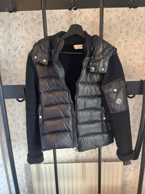 Svart pufferjacka från Moncler - Snygg svart pufferjacka från Moncler med stickade ärmar och huva. Jackan har en quiltad front i glansigt material, dragkedja och fickor med tryckknappar. På vänster ärm finns en ficka med Moncler-logga. Perfekt för kalla dagar.