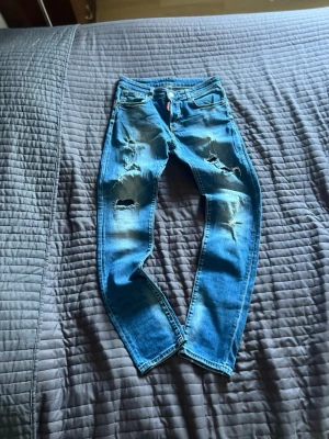 Dsquared2 - Säljer ett par blå jeans med flera slitningar och hål på både fram- och baksida. Jeansen har en klassisk femficksmodell och är tillverkade i denim med en tvättad look. Perfekta för en avslappnad och trendig stil.