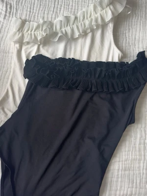 Svart och vit body med volang - Två snygga bodys från H&M, en svart och en vit, båda med en axel och bred volangdetalj längs överkanten. Tillverkade av 87% återvunnen polyester och 13% elastan för en stretchig och bekväm passform. 