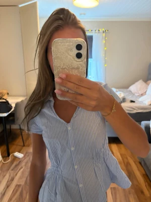 Blåvit randig kort klänning - Säljer en söt blåvit randig kort klänning med knappar framtill och v-ringad hals. Klänningen har korta ärmar och markerad midja för en feminin siluett. Perfekt för varma dagar och enkel att styla med sneakers eller sandaler.