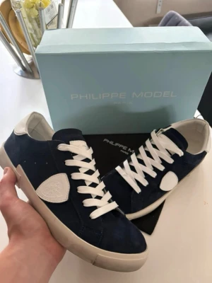 Philippe Model blåa sneakers - Snygga sneakers från Philippe Model i mörkblå mocka med vita detaljer och vita skosnören. Skorna har en klassisk låg siluett och vit sula. Perfekta för dig som gillar stilrena och trendiga sneakers med premiumkänsla. Kontakta mig via funderingar😊
