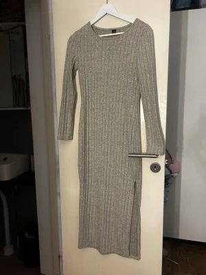 Beige ribbad långklänning - Långärmad beige ribbad långklänning med rund hals och slits på sidan. Klänningen har en kroppsnära passform och är tillverkad i ett mjukt stickat material som passar perfekt för kyligare dagar. Aldrig använd, storlek L passar även M