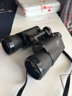 Svart kikare 7x50 med rem - Säljer ett par klassiska svarta kikare med 7x50 förstoring och Field 7°. Kikarna har robust design i metall och plast med räfflat grepp och justerbar fokus. Perfekt för fågelskådning eller utflykter. Snygg och tidlös look med svart rem för enkel transport.