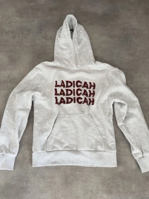 Grå hoodie med LADICAH-tryck - Snygg ljusgrå hoodie från LADICAH med stor huva och känguruficka. Framsidan har tre röda LADICAH-tryck med vita detaljer och ärmen har en broderad logga. Perfekt för en chill och trendig look.