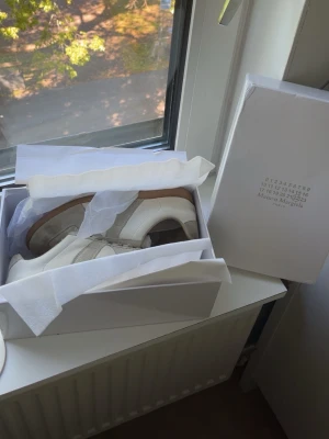 Maison Margiela Gats vita sneakers - Skorna är köpta här på plick och är andvända. Sista bilden ser man än liten defekt som syns nära, men inte så synlig under andvändning. Det är ett par klassiska vita sneakers från Maison Margiela med beige mockadetaljer. Perfekta för dig som gillar minimalistisk och trendig stil. Har du frågor/ funderingar hör av dig 🤝😊