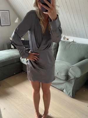 Grå omlottklänning med lång ärm - Snygg grå omlottklänning med lång ärm och v-ringad front. Klänningen har en kort längd och en elegant, enkel design som ger en stilren look. Tillverkad i ett glansigt material som ger en lyxig känsla.