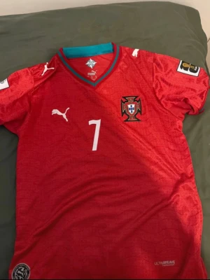 Portugal fotbollströja Ronaldo #7 Puma  - Röd Portugal fotbollströja från Puma med nummer 7 och Ronaldo på ryggen. Tröjan har korta ärmar, broderad landslagssköld och vita Puma-loggor. Turkos detalj vid kragen och tryck på ärmarna. Perfekt för dig som älskar fotboll och Ronaldo.