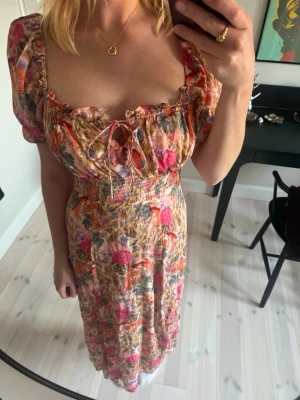 Blommig långklänning med snörning - Superfin långklänning med blommigt mönster i rosa, rött och beige toner. Klänningen har puffärmar, smock i midjan och söt snörning vid bysten.