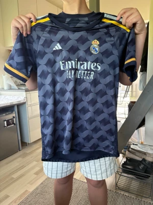 Real Madrid matchtröja Vini Jr #7 - Snygg mörkblå Real Madrid matchtröja från Adidas med Vini Jr och nummer 7 på ryggen. Tröjan har gula detaljer på ärmarna och axlarna, samt klubbmärke och sponsortryck på bröstet. Tillverkad i lätt och ventilerande material, perfekt för fotbollsträning eller match.