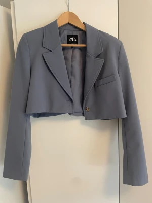 Gråblå cropped kavaj från Zara - Stilren grå cropped kavaj från Zara med klassisk krage, lång ärm och knappdetalj framtill. Kavajen har en bröstficka och är helfodrad för extra komfort. Perfekt för en trendig och modern look.