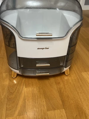 Sminkförvaringsbox med lådor - Praktisk sminkförvaringsbox i vit och grå plast med transparent lock och flera utdragbara lådor. Boxen har guldfärgade handtag och ben, samt texten 'storage box' på framsidan. Perfekt för att organisera sminkborstar, hudvård och andra skönhetsprodukter.