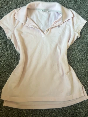 Ljusrosa pikétopp från Tommy Hilfiger - Snygg ljusrosa pikétopp från Tommy Hilfiger med klassisk krage, korta ärmar och liten broderad logga på bröstet. Tillverkad i mjuk bomull som känns skön mot huden. Perfekt till jeans eller kjol för en clean och fräsch look.