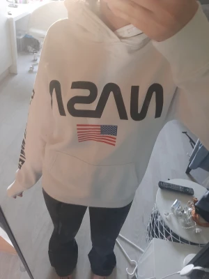 Vit NASA hoodie med tryck - Säljer en vit hoodie med stor svart NASA-logga på bröstet och amerikansk flagga under. Tryck i svart även längs ena ärmen. Klassisk känguruficka framtill och huva. Perfekt för dig som gillar rymdtema och streetwear. Storleken är 158/164, alltså s eller xs.