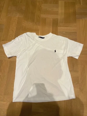 Vit t-shirt från Ralph Lauren - Klassisk vit t-shirt från Ralph Lauren med den ikoniska lilla loggan broderad på bröstet. T-shirten har rund hals och korta ärmar, perfekt basic-plagg i garderoben. Tillverkad i mjuk bomull för skön känsla.