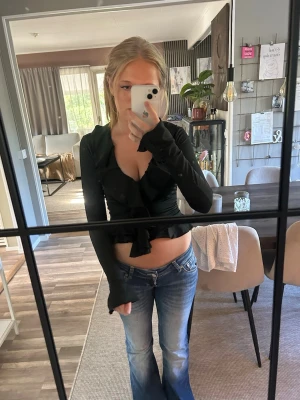 Svart omlottopp med volang - Trendig svart omlottopp med djup v-ringning och volangdetaljer längs kanten. Toppen är croppad och har långa ärmar som ger en snygg och modern look. Perfekt att styla med jeans eller kjol för en cool outfit.
