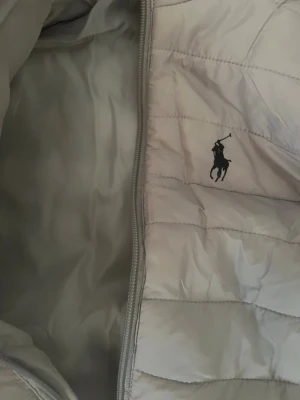 Ljusgrå dunväst från Polo Ralph Lauren - Snygg ljusgrå dunväst från Polo Ralph Lauren med klassisk broderad logga på bröstet. Västen har quiltad design, dragkedja framtill och är tillverkad i ett glansigt material som ger en modern look. Perfekt att slänga över en hoodie eller tröja för extra värme.