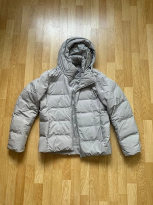 Ljusgrå pufferjacka från Uniqlo - Säljer en ljusgrå pufferjacka från Uniqlo med huva och dragkedja. Jackan är lätt, vindtät och har vattenavvisande material. Perfekt för kalla dagar och har en stilren, boxig passform som är enkel att matcha med olika outfits.