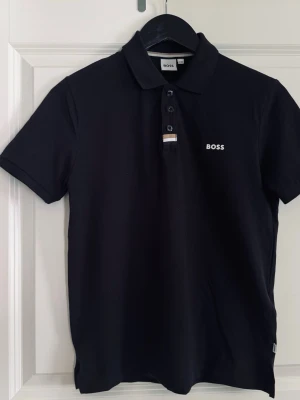 BOSS Polo Shirt Black – 12Y / XS 🖤 - Säljer en jättefin BOSS pikétröja i mörksvart färg i storlek 12 år / XS. Färgen är helsvart i verkligheten (kan se annorlunda ut på bilderna på grund av ljus/kamera). En stilren och tidlös modell med fina detaljer som ger en mer exklusiv känsla. Har BOSS-knappar och logga framtill. Loggan är i fint skick och inte sprucken eller sliten. Passar perfekt både till vardag och en mer uppklädd look och är enkel att matcha med det mesta 🖤 Inga hål, fläckar eller liknande – mycket fint skick.