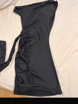Svart oneshoulder långklänning - Snygg svart långklänning med oneshoulder-design och draperad effekt längs sidan. Klänningen har en lång slits och är tillverkad i ett mjukt, stretchigt syntetmaterial som sitter tight och markerar figuren. Perfekt för dig som vill sticka ut med en elegant och modern look.