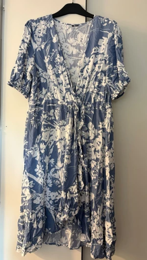 Blåvit blommig omlottklänning - Söt blåvit kortklänning med blommigt mönster och omlottmodell. Klänningen har v-ringning, korta ärmar med resår och ett knytband i midjan. Tillverkad i ett lätt och luftigt material, perfekt för varma dagar.
