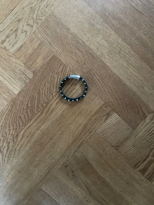 Svart armband med pärldetaljer - Snyggt svart armband med små pärlor och flätad design. Armbandet har ett stilrent magnetlås i silverfärg och passar perfekt för dig som gillar en cool och modern look. Materialet är troligtvis konstläder och metall.